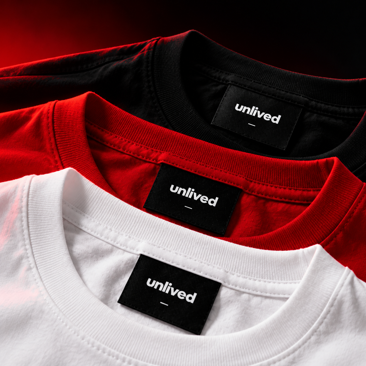 Détail des cols et étiquettes UNLIVED des trois t-shirts noir, rouge et blanc empilés